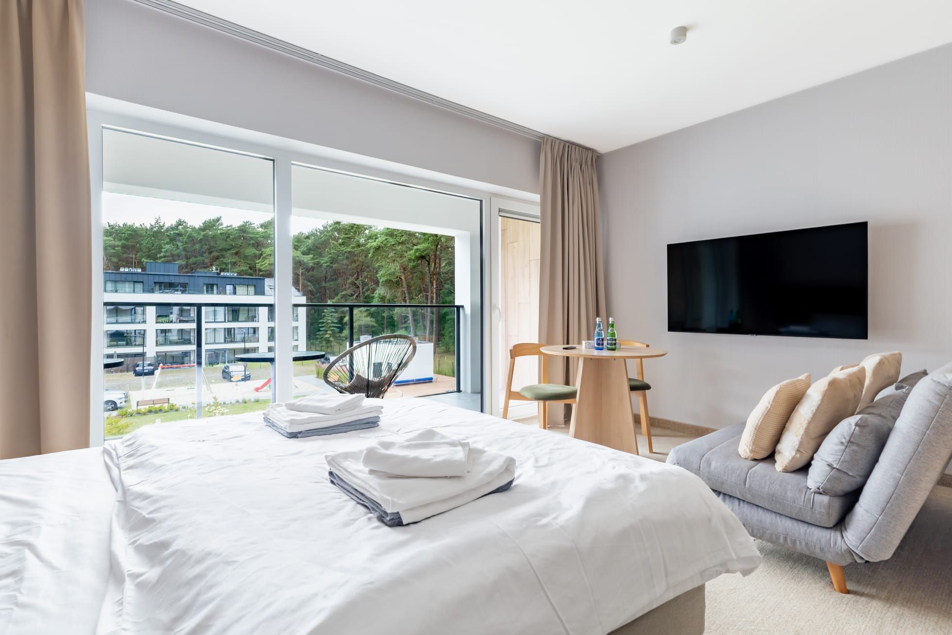 Habitación de hotel con cama, sofá, televisor y balcón con vistas a edificios y árboles.