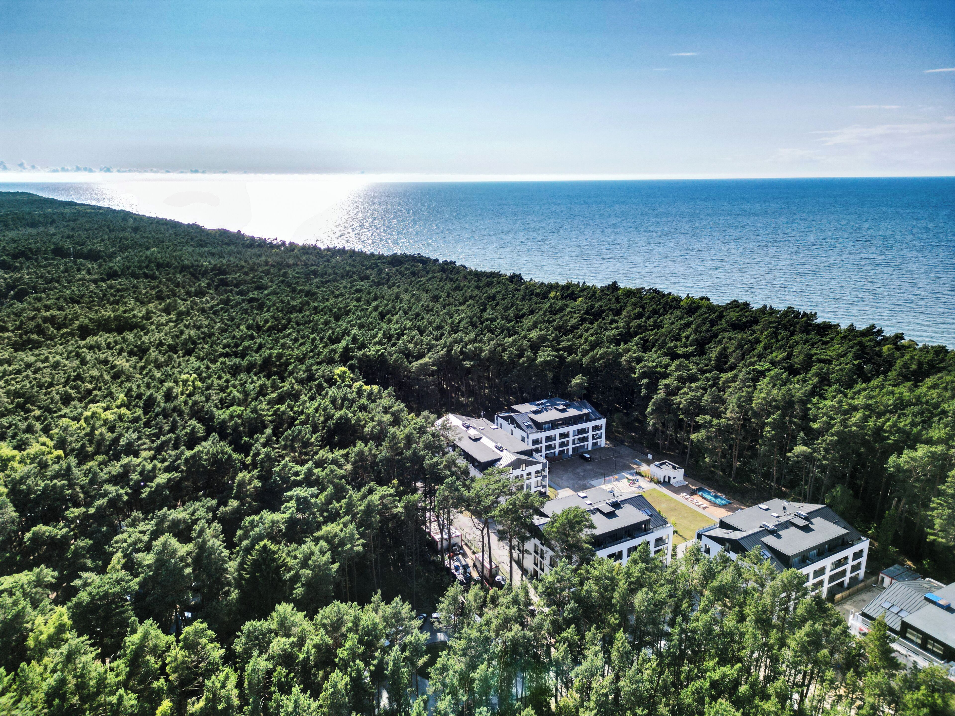 An aerial view of a forested area with a modern building complex featuring multiple buildings, a parking area, and a view of the sea.
