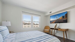 A hotel room with a bed, a desk, a chair, a TV, and a window with a view of buildings. - Immaculant One Bedroom Condo, Conveniently Located On 40th ST (Ocean City)