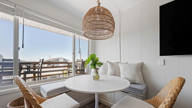 A room with a round white table, wicker chairs, a hanging rattan light fixture, a white sofa with cushions, and a large window offering a view of buildings and a balcony. - Immaculant One Bedroom Condo, Conveniently Located On 40th ST (Ocean City)