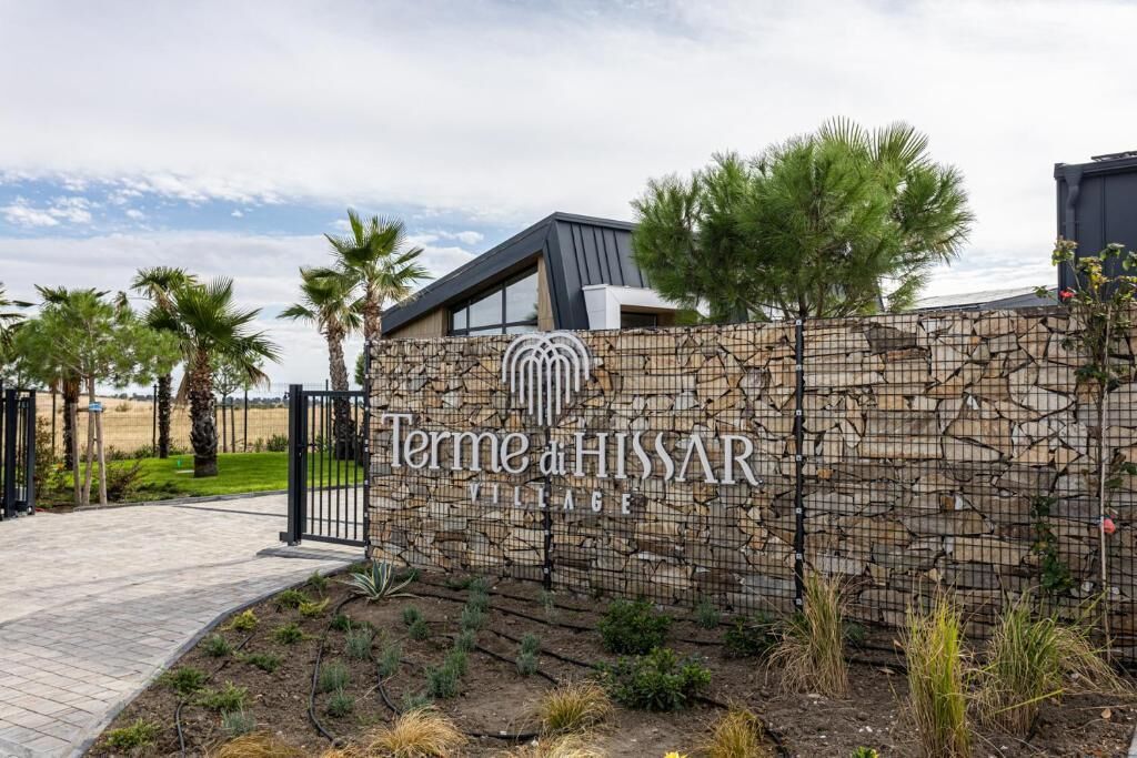 Un muro de piedra con el texto 'Terme di HISSAR VILLAGE' y un edificio moderno con techo inclinado.