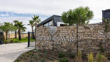 Un muro de piedra con el texto 'Terme di HISSAR VILLAGE' y un edificio moderno con techo inclinado.