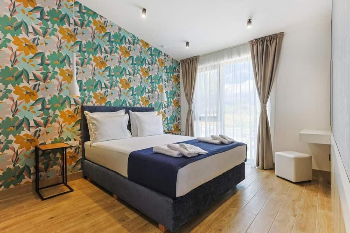 Habitación de hotel con cama, mesita de noche, pequeño taburete, ventana grande con cortinas y papel tapiz floral.