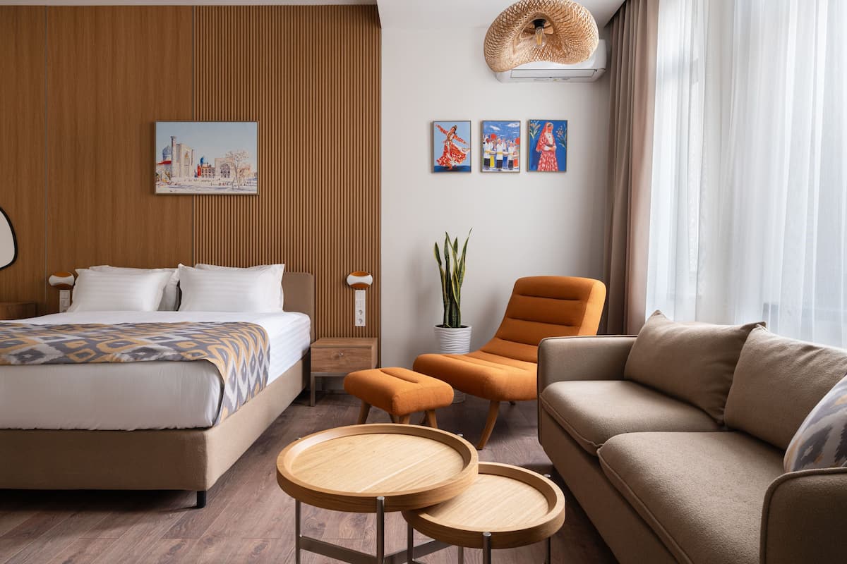 A hotel room with a bed, a sofa, two side tables, a potted plant, and artwork on the wall.