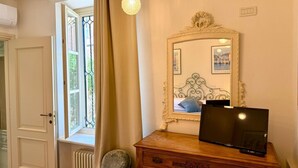 Ein Zimmer mit einem Holzkoffer, einem Fernseher darauf, einem Spiegel mit Zierrahmen und einem Fenster mit Vorhängen.
