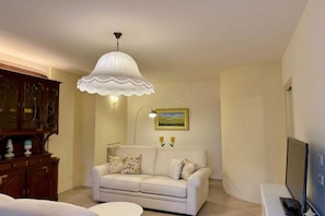 A living room with a white sofa, a white table, a TV, a lamp, and a painting on the wall.