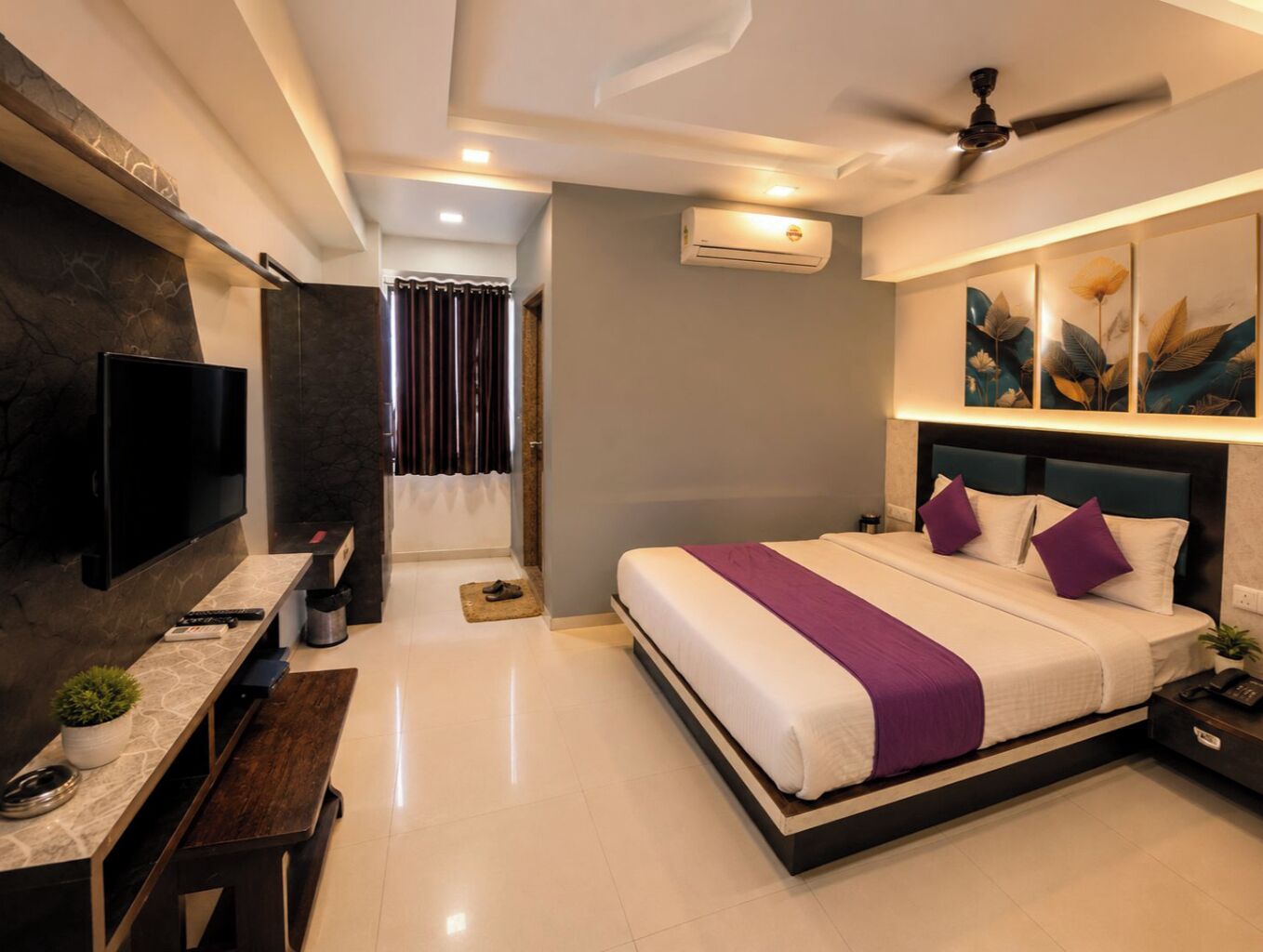 A modern hotel room with a large bed, a desk with a flat-screen TV, a ceiling fan, and artwork on the walls.