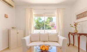 Ein Zimmer mit einem weißen Sofa, einem Holztisch mit Obstschale, einem Schrank und einem Fenster mit Vorhängen.