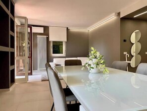 A modern dining area with a glass table, white chairs, a vase of flowers, and a wall-mounted mirror. - Elegant modern apartment with pool and panoramic balconies (Stresa)