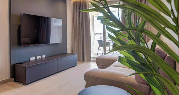 A modern living room with a flat-screen TV mounted on the wall, a dark console table, a beige sofa, and a large green plant. - A Stylish 2BR seafront home in Qawra w/WIFI & AC (San Pawl il-Baħar)