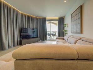 A modern living room with a large sofa, a flat-screen TV, and a balcony with a table and chairs. - Seafront Seafront 3BR home in Qawra w/WIFI & AC (San Pawl il-Baħar)