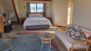 A bedroom with a bed, a sofa, a small table, a dresser, a fan, and a window with curtains.