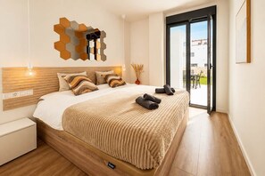 Ein modernes Schlafzimmer mit einem Holzkopfteil, einem großen Bett mit einer beigefarbenen Decke, einem hexagonalen Spiegel an der Wand und einer Glastür, die zu einem Außenbereich führt.