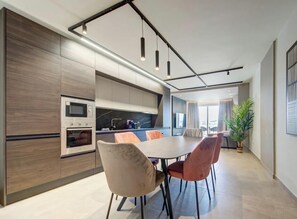 A modern kitchen with built-in appliances, a dining area with a round table and chairs, and a large window providing natural light. - Exceptional 2BR seafront home in Qawra w/WIFI & AC (San Pawl il-Baħar)