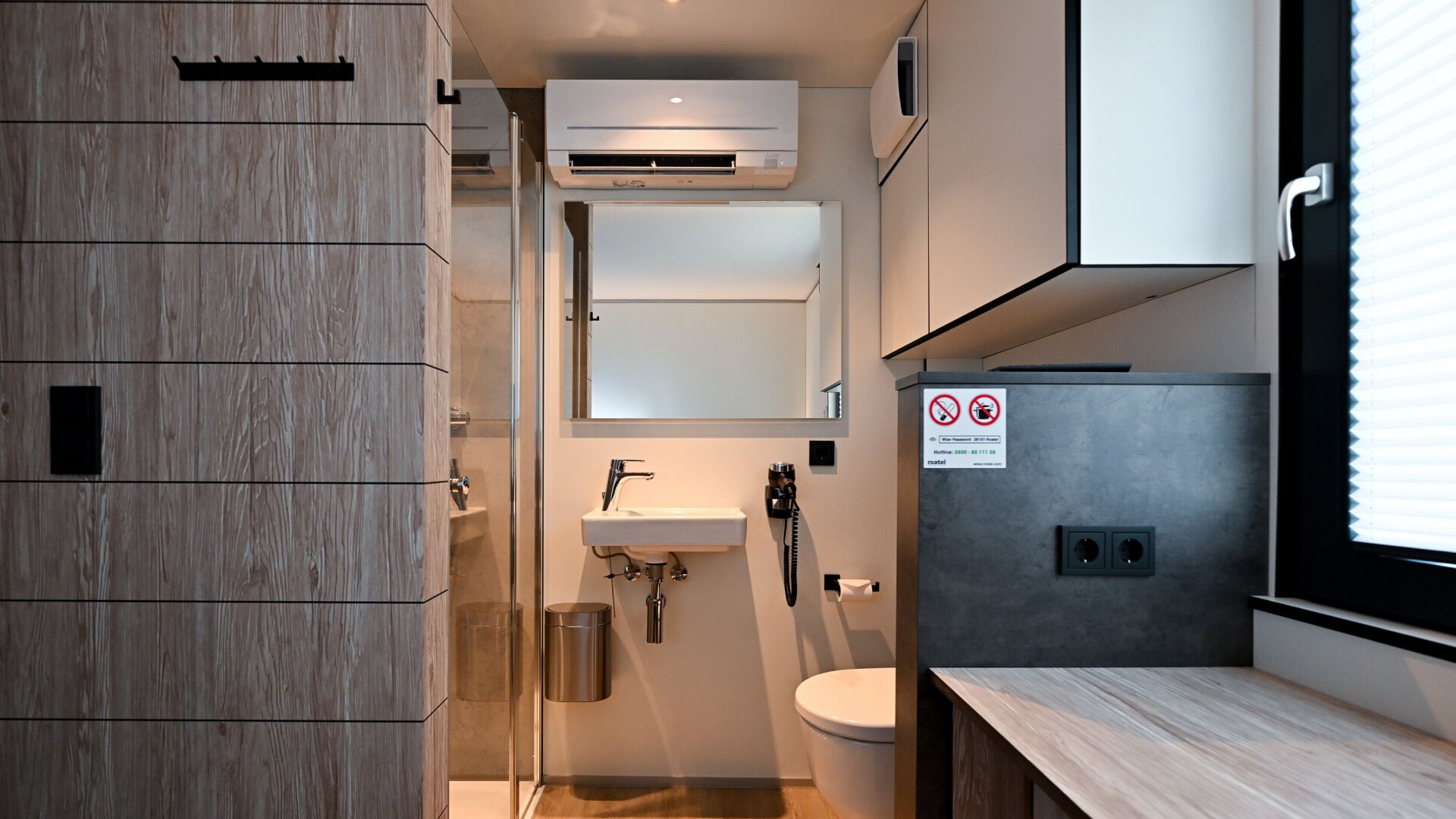 A modern bathroom with a shower, sink, and toilet. There is a towel rack, a mirror, and a sign indicating no smoking.