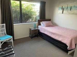Un dormitorio con cama, una silla, una mesita de noche, una ventana con cortinas y un cuadro en la pared.