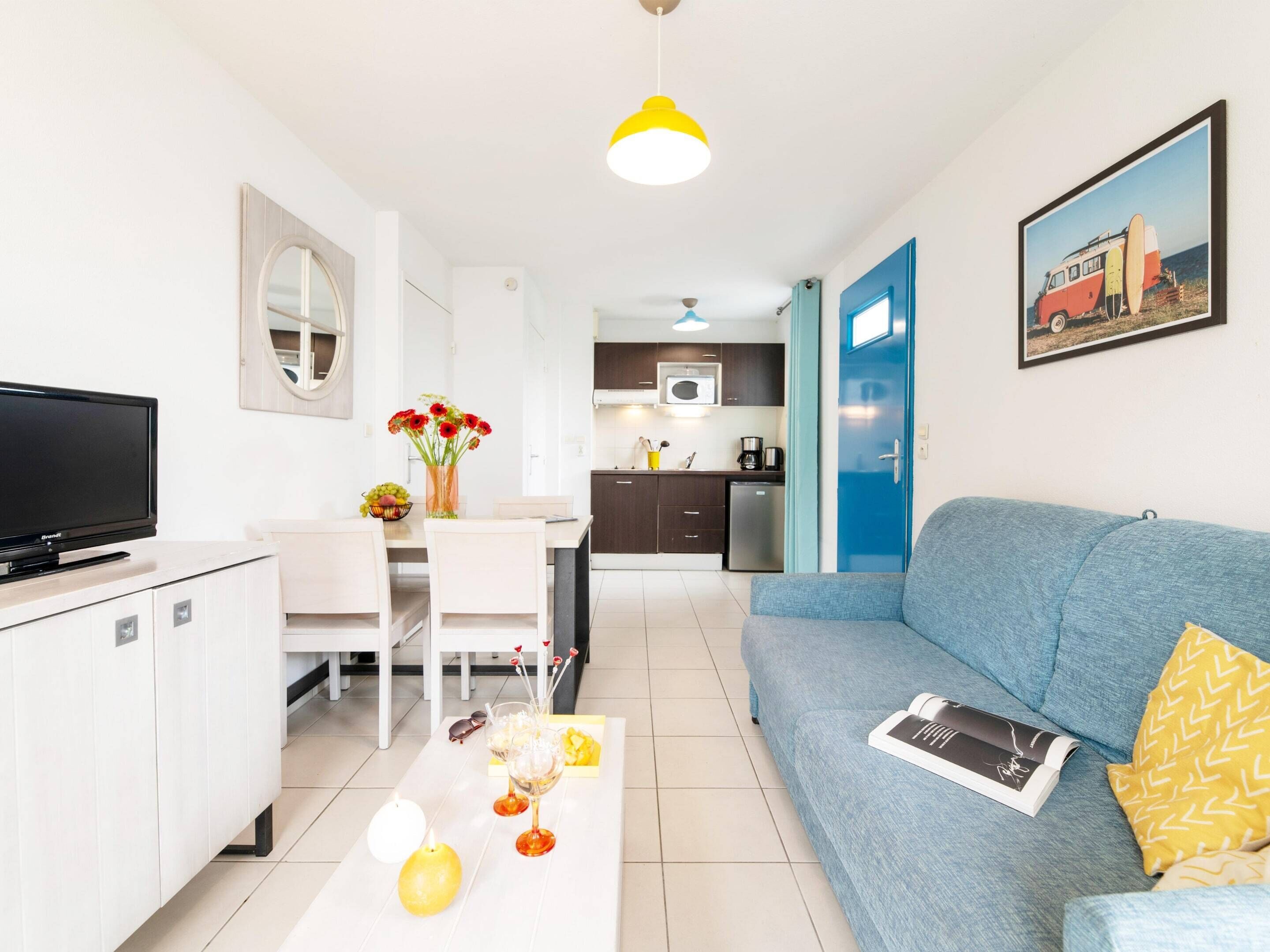 Una sala de estar moderna con un televisor de pantalla plana, una mesa de comedor con sillas, un sofá, una mesa de centro con objetos decorativos y una cocina al fondo.