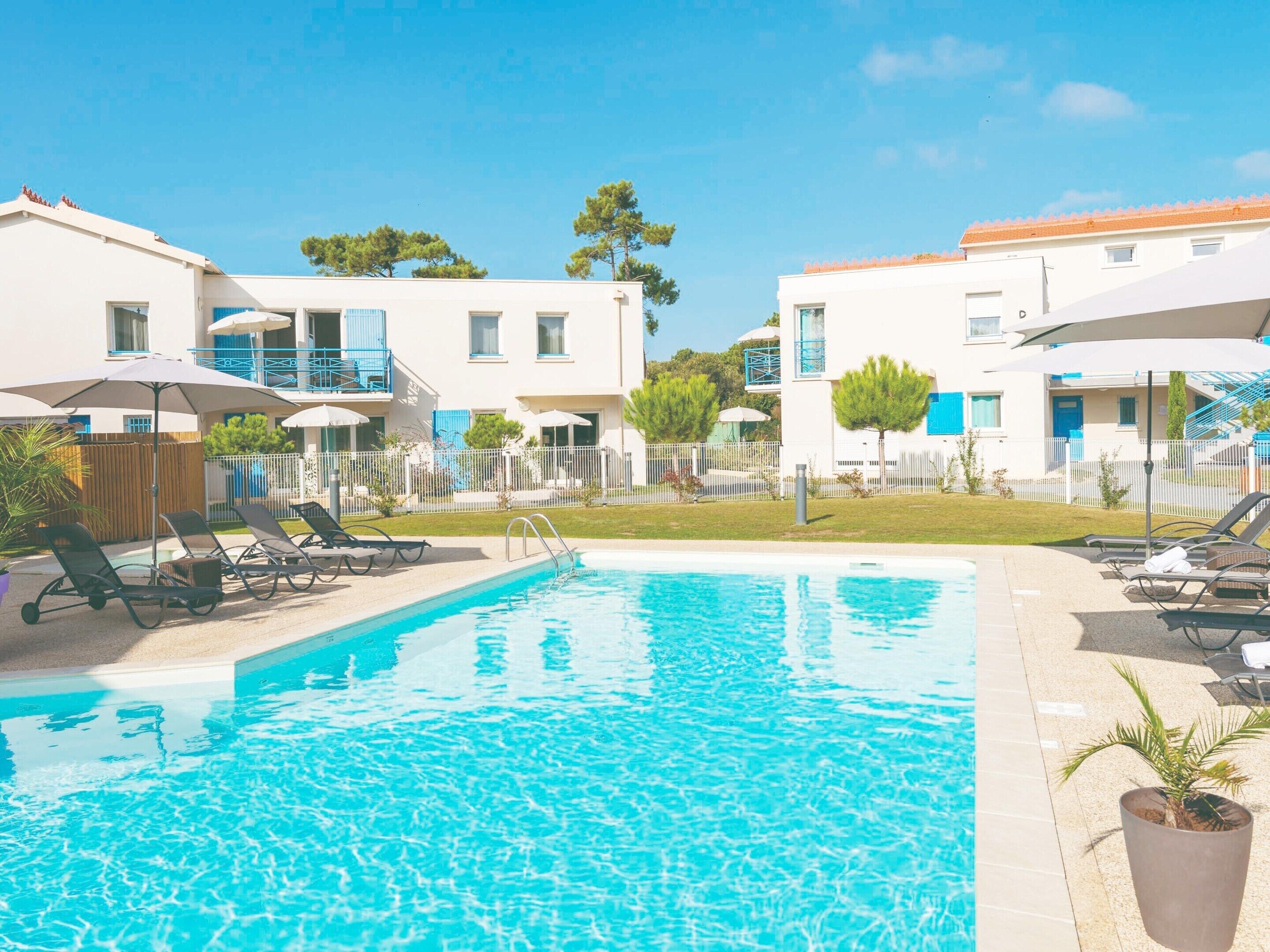Une piscine avec des chaises longues, un espace délimité par une clôture et des bâtiments dotés de balcons et de fenêtres.