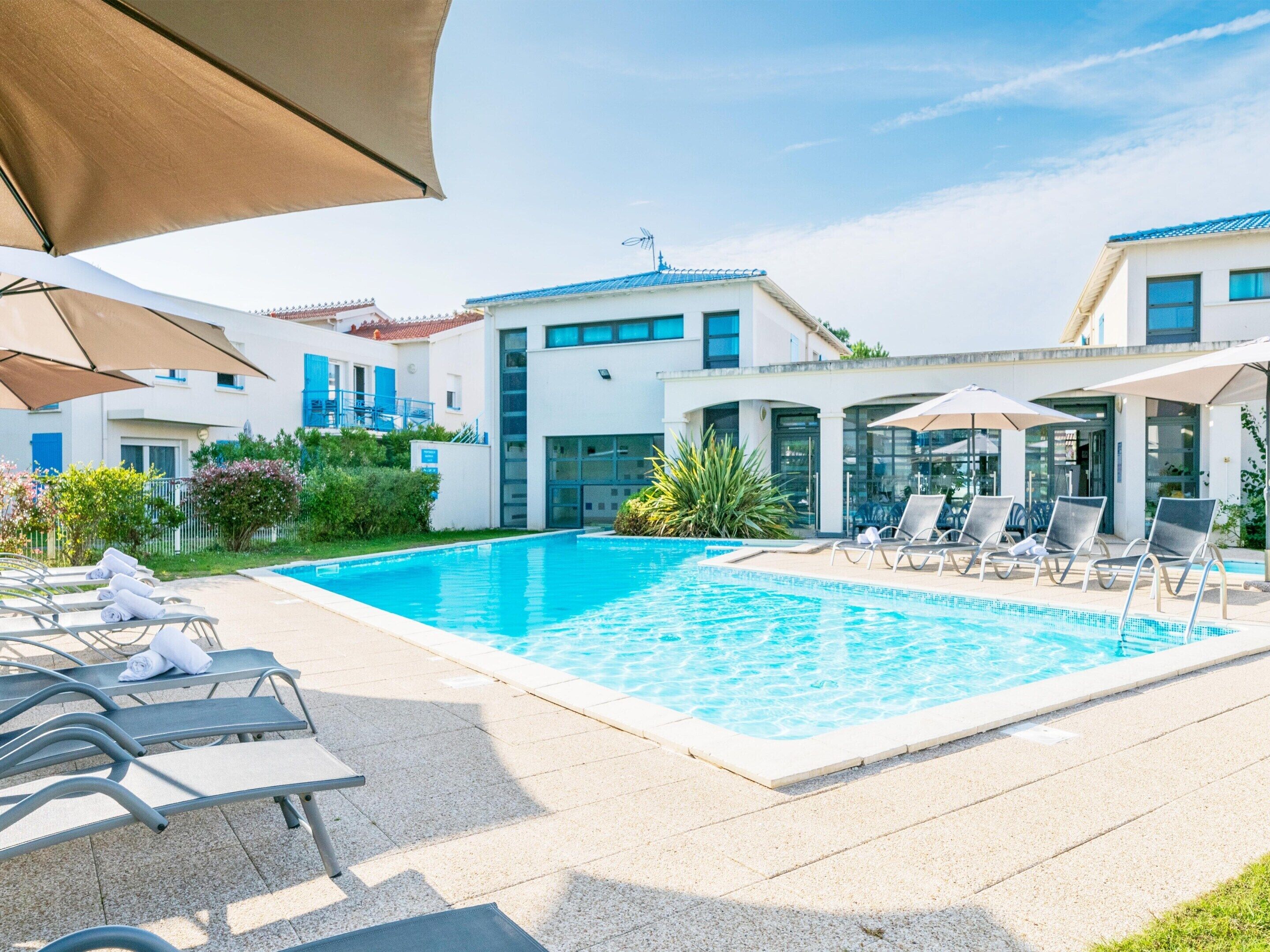 A swimming pool with lounge chairs and umbrellas, surrounded by buildings with large windows and a clear sky.