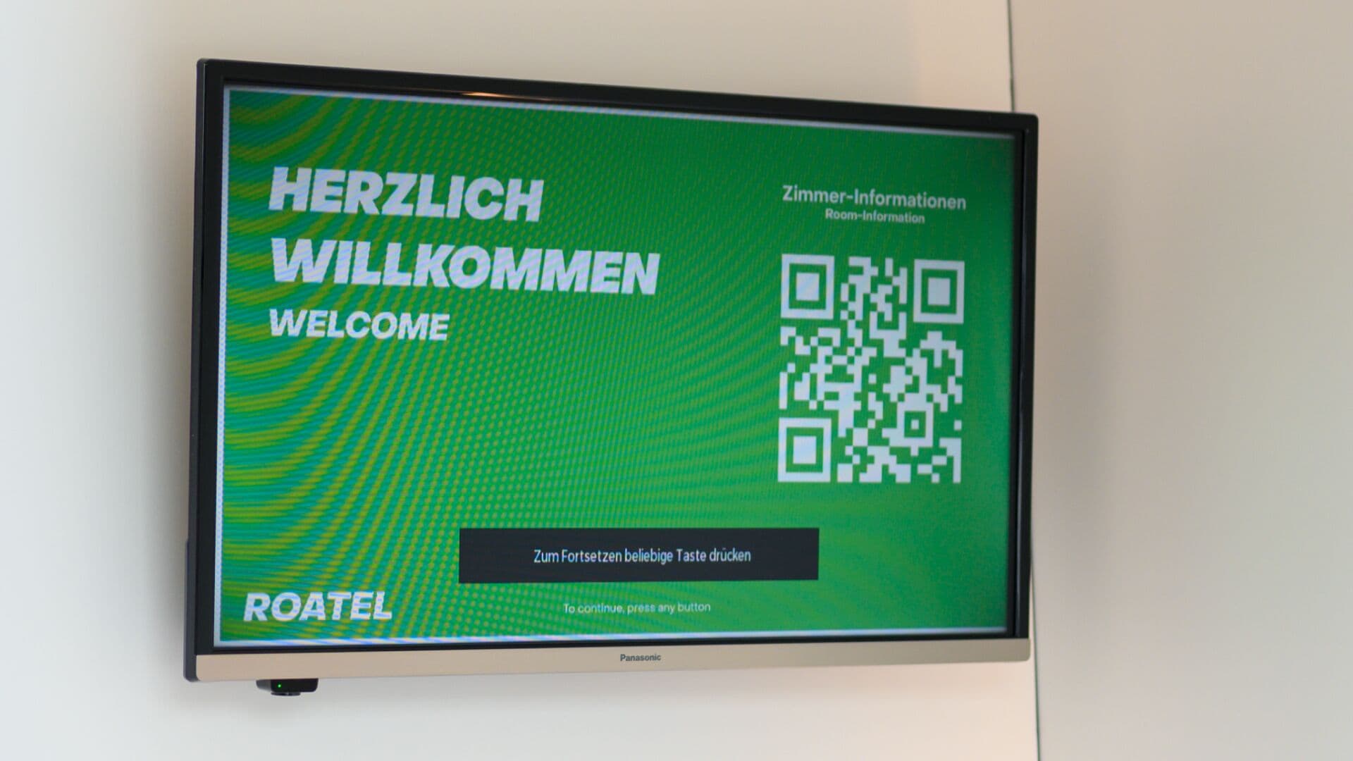 Ein digitaler Begrüßungsbildschirm mit einem QR-Code und Text auf Deutsch, der eine Willkommensbotschaft und möglicherweise Informationen zum Hotel oder zum Ort anzeigt.