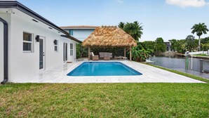 Une piscine dotée d’une structure de toit en chaume, un bâtiment blanc et un élément décoratif aquatique.