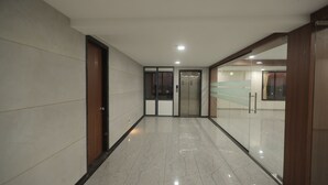 Um corredor com piso de mármore polido, portas de madeira e um elevador de vidro.