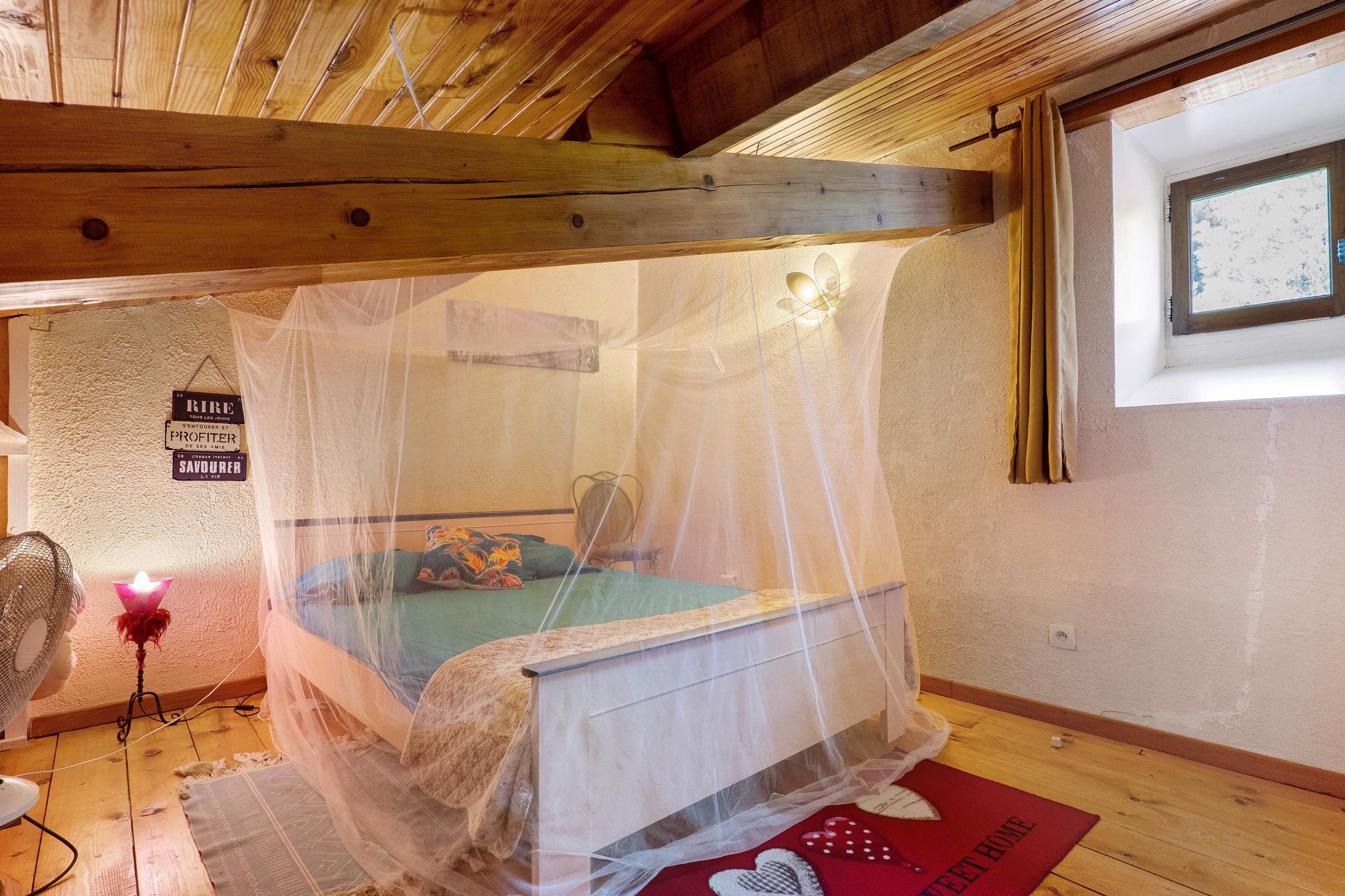 Ein Zimmer mit einem Bett unter einem Moskitonetz, einem kleinen Tisch mit einer Lampe und einem Fenster mit Vorhängen.