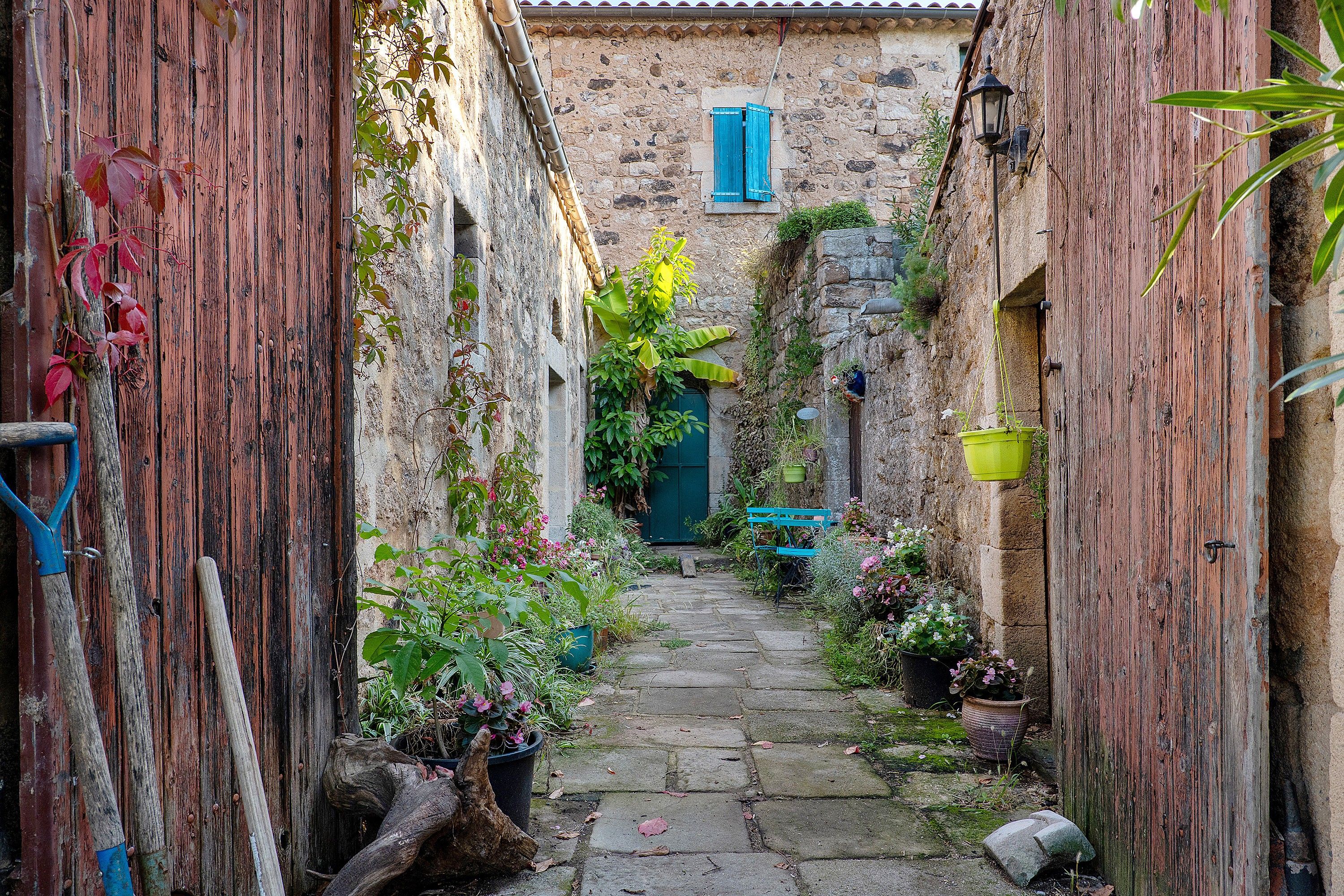 A narrow stone pathway flanked by rustic wooden walls, leading to a building with a blue shuttered window.