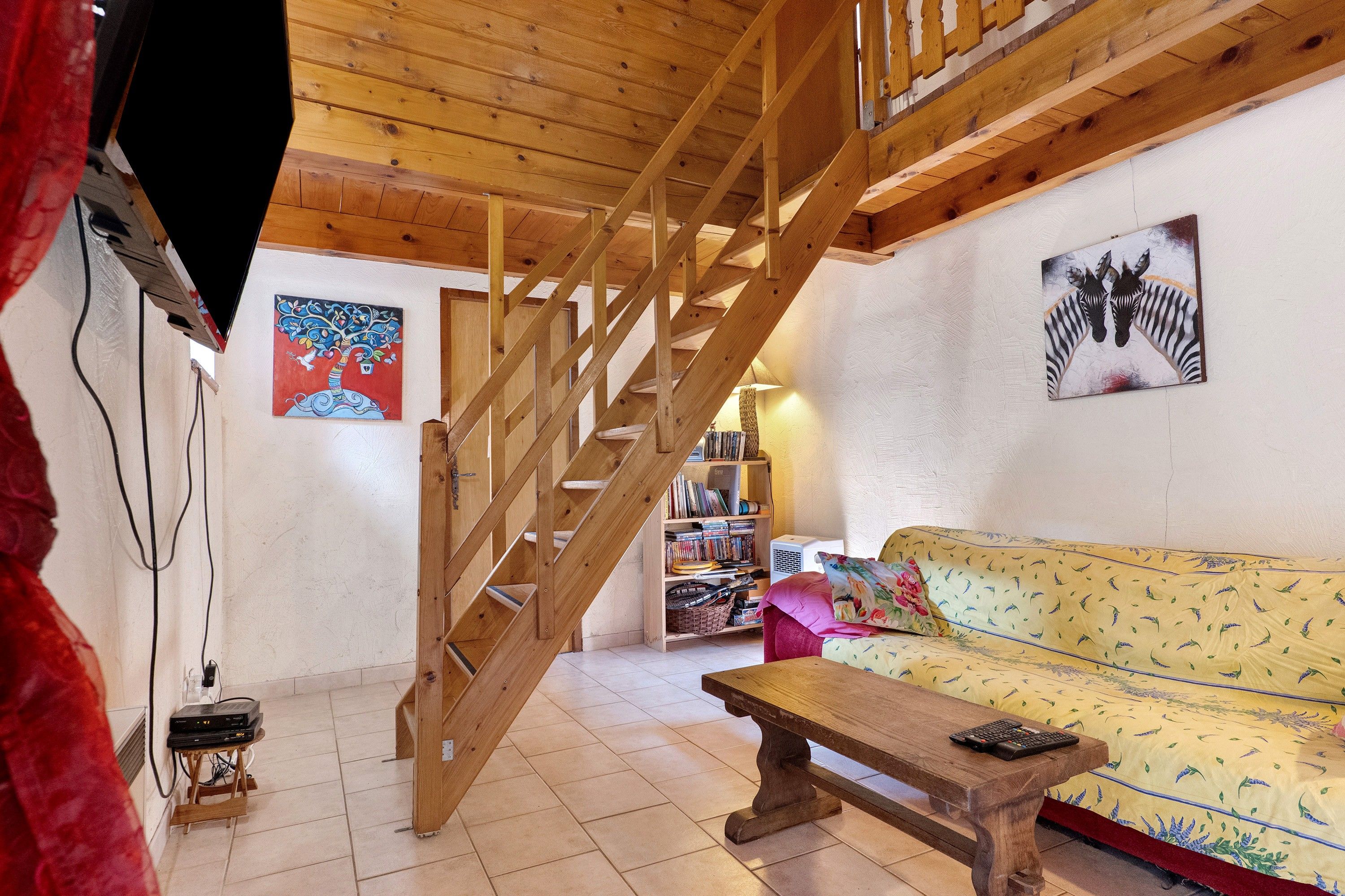 A wooden staircase leading to an upper level, a sofa with a patterned cover, a wooden table, and a bookshelf filled with books.