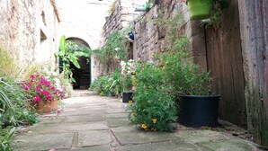 A narrow stone pathway leading to a rustic building with a wooden door, surrounded by lush greenery and potted plants. - Holiday Home 'La Fée Du Val - Cottage in Ardèche' with Private Pool, Wi-Fi and Air Conditioning (Rosières)