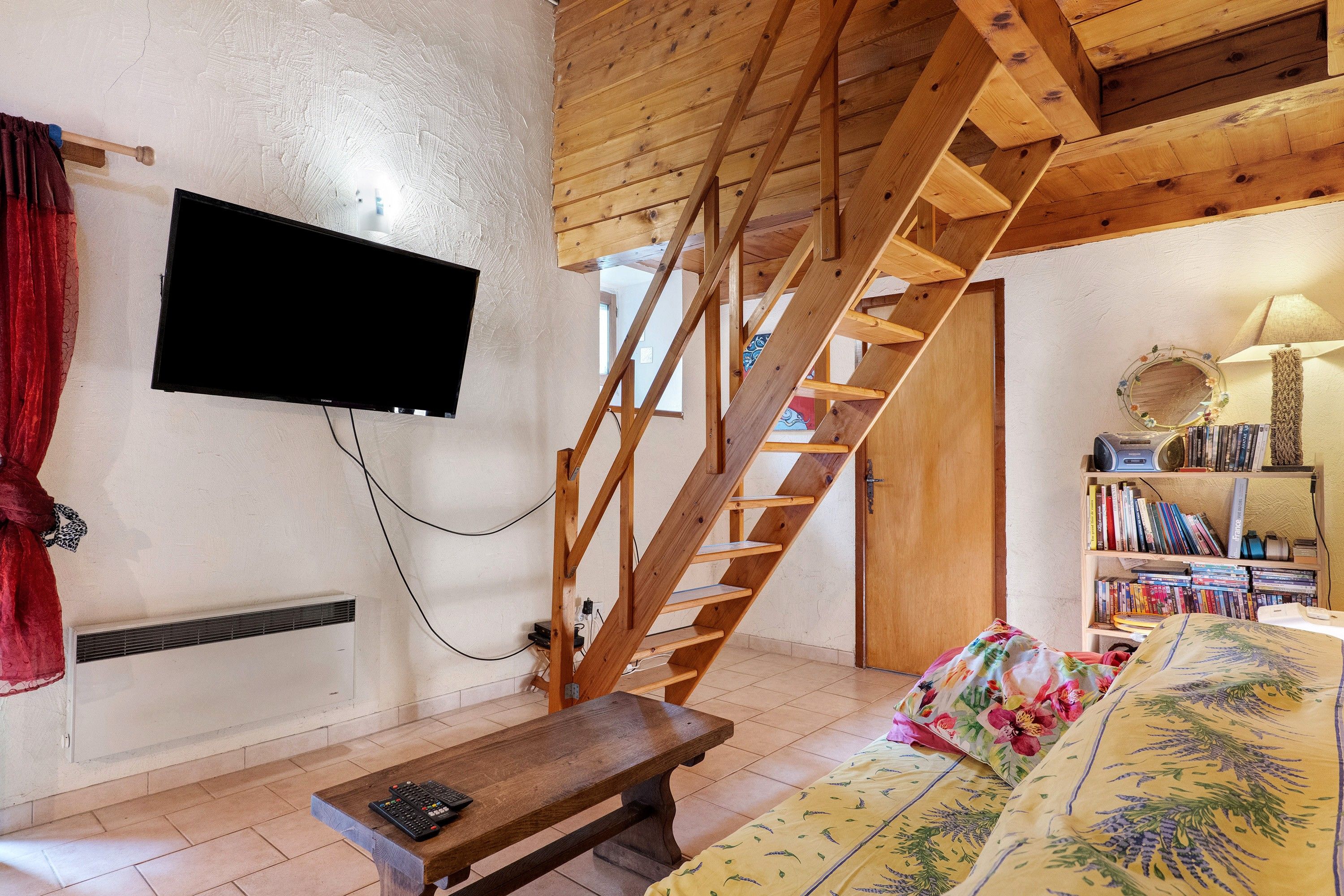 A wooden staircase leading to an upper level, a flat-screen TV mounted on the wall, a bookshelf filled with books, and a bench in the foreground.
