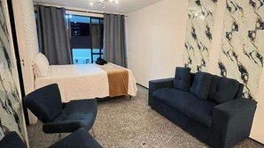 A hotel room with a bed, a sofa, two armchairs, a carpeted floor, a window with curtains, and a wall with a marble-like pattern. - Atlantico 204 apartment, 50m2, 1 bedroom, sleeps up to 4 people. (Fortaleza)