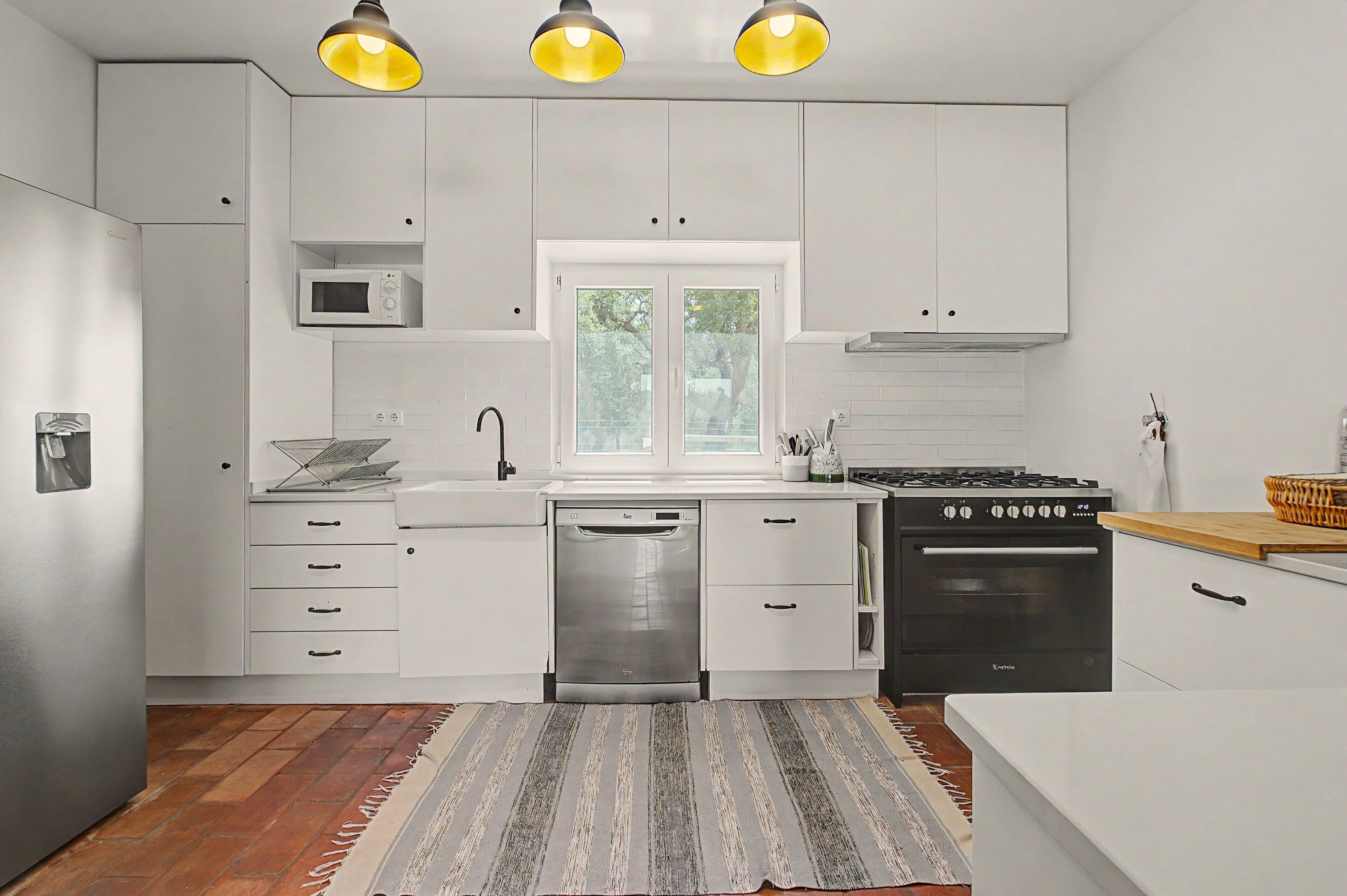 A modern kitchen with white cabinets, a dishwasher, a stove, and a window with a view of greenery.