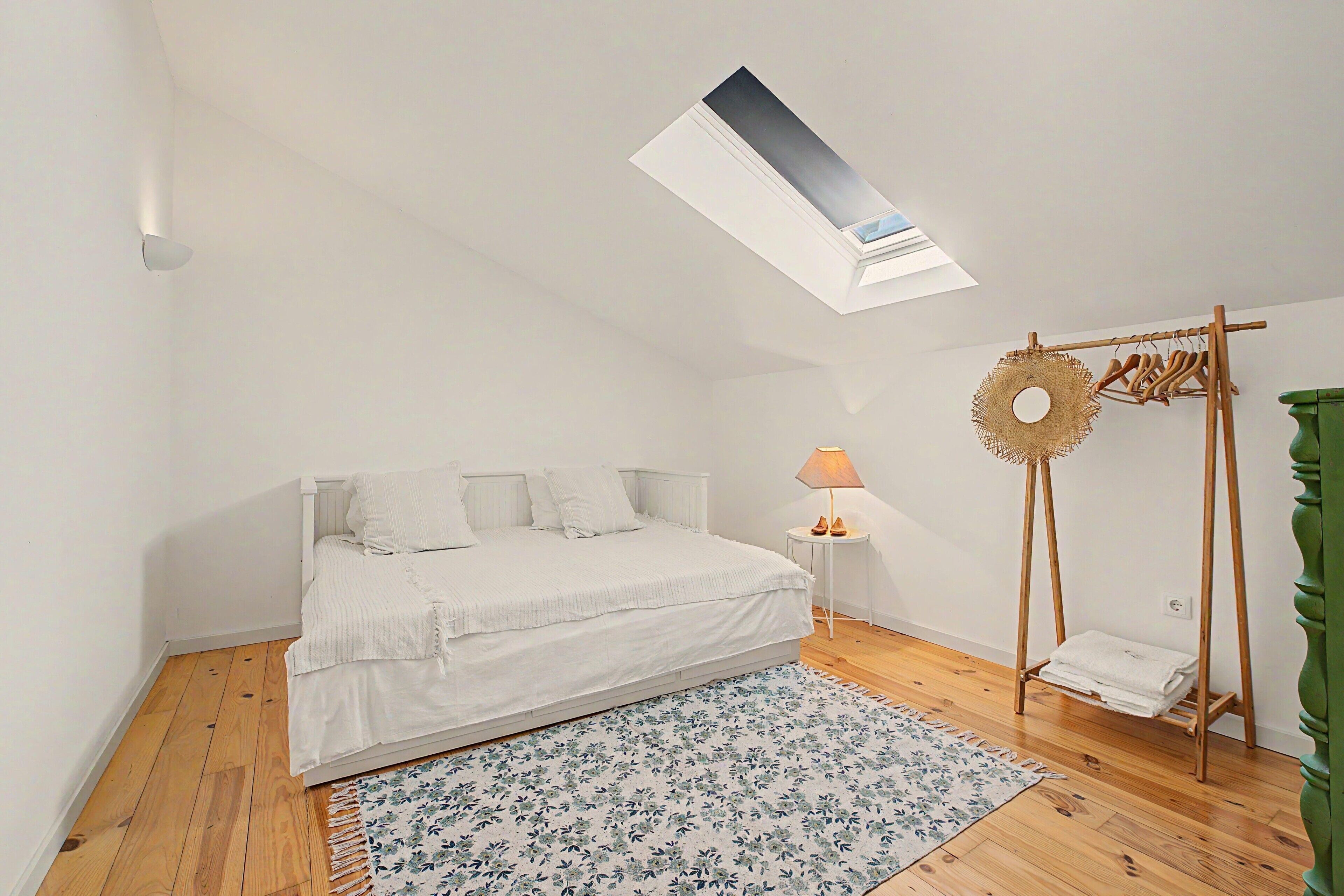 A bedroom with a skylight, a bed, a small bedside table, a lamp, a hanging rack, and a green plant.