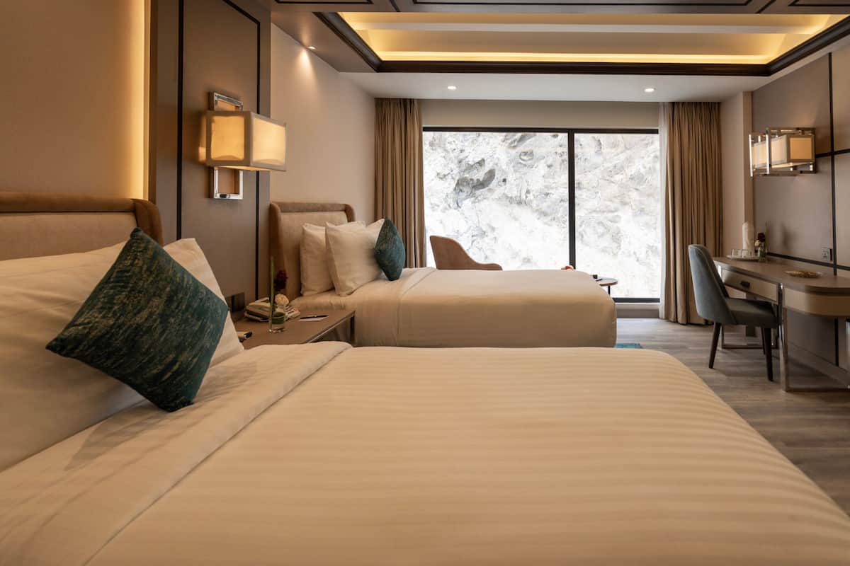 Habitación de hotel con dos camas, un escritorio, una silla y un amplio ventanal con cortinas.