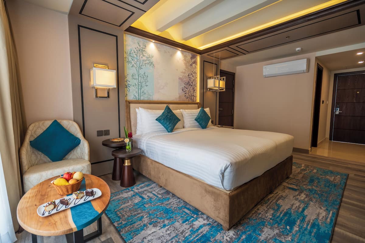 Habitación de hotel con cama, una silla, una mesita con una cesta y un plato de comida, y una pared con un panel decorativo.
