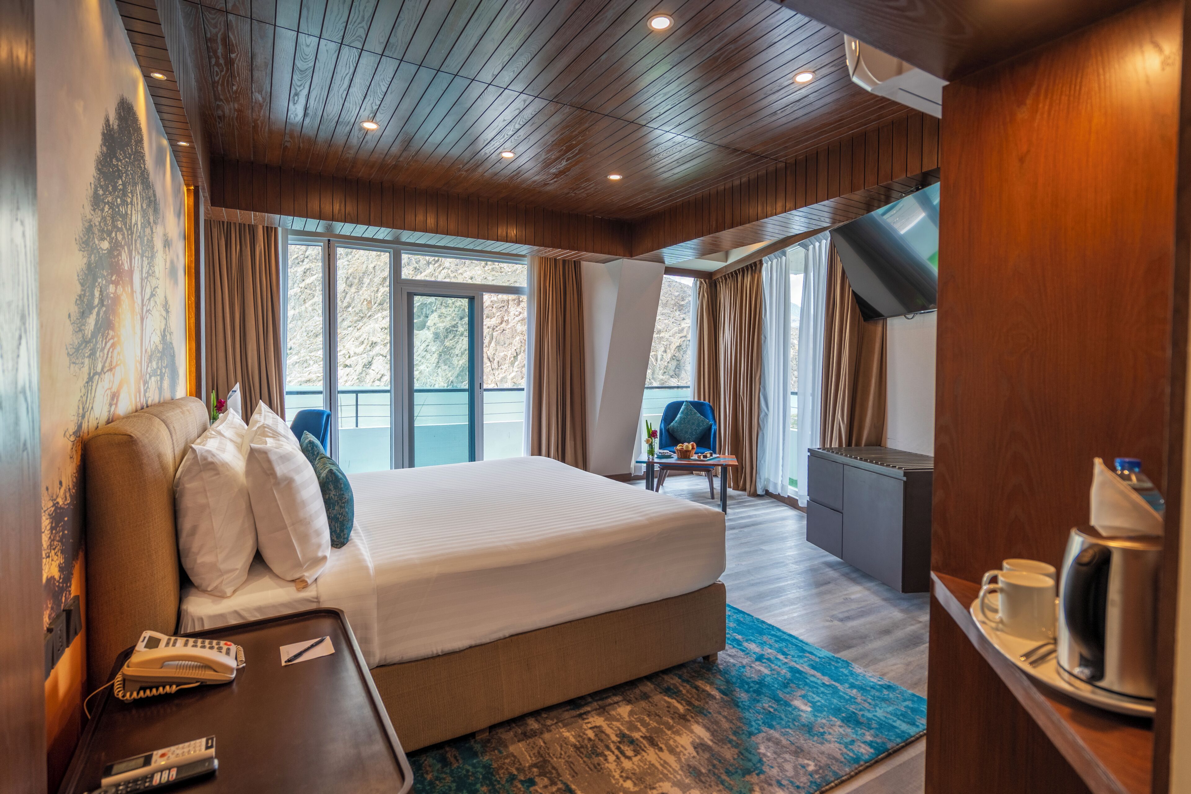Quarto de hotel com uma cama grande, uma escrivaninha com telefone e controles remotos, uma cafeteira e vista para o exterior através de janelas amplas.