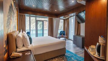 Quarto de hotel com uma cama grande, uma escrivaninha com telefone e controles remotos, uma cafeteira e vista para o exterior através de janelas amplas.