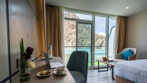 A hotel room with a bed, a desk with a laptop, a chair, a small table with a bowl of fruit, and a view of a mountainous landscape through large windows. - Pearl Continental Attabad Lake (Aina Abad)