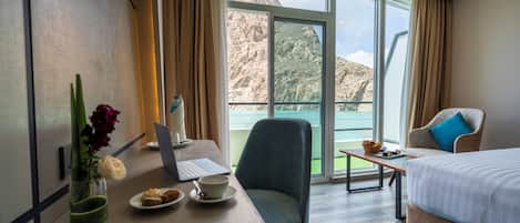 Habitación de hotel con cama, escritorio con computadora portátil, silla, mesita con frutero, y vista a un paisaje montañoso a través de amplias ventanas.