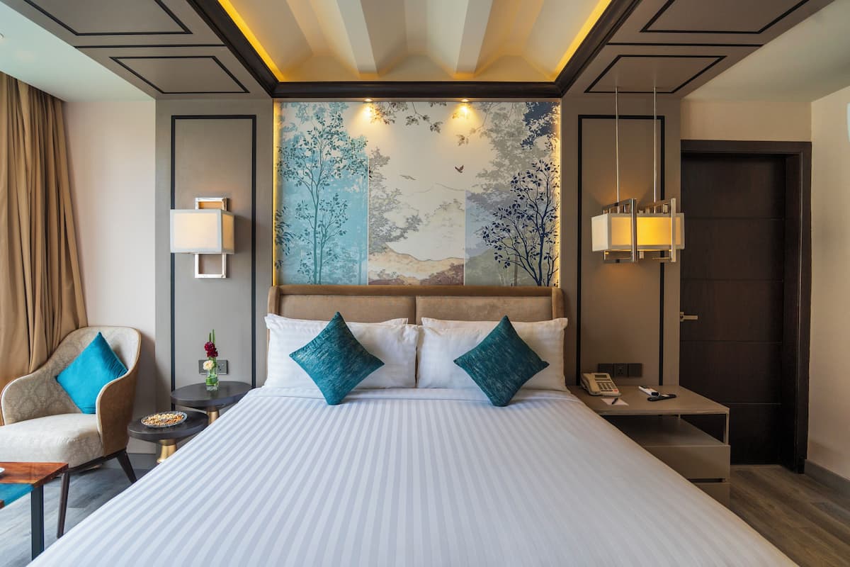 Habitación de hotel con una cama grande, una silla, una mesita con una lámpara y un cuadro en la pared.