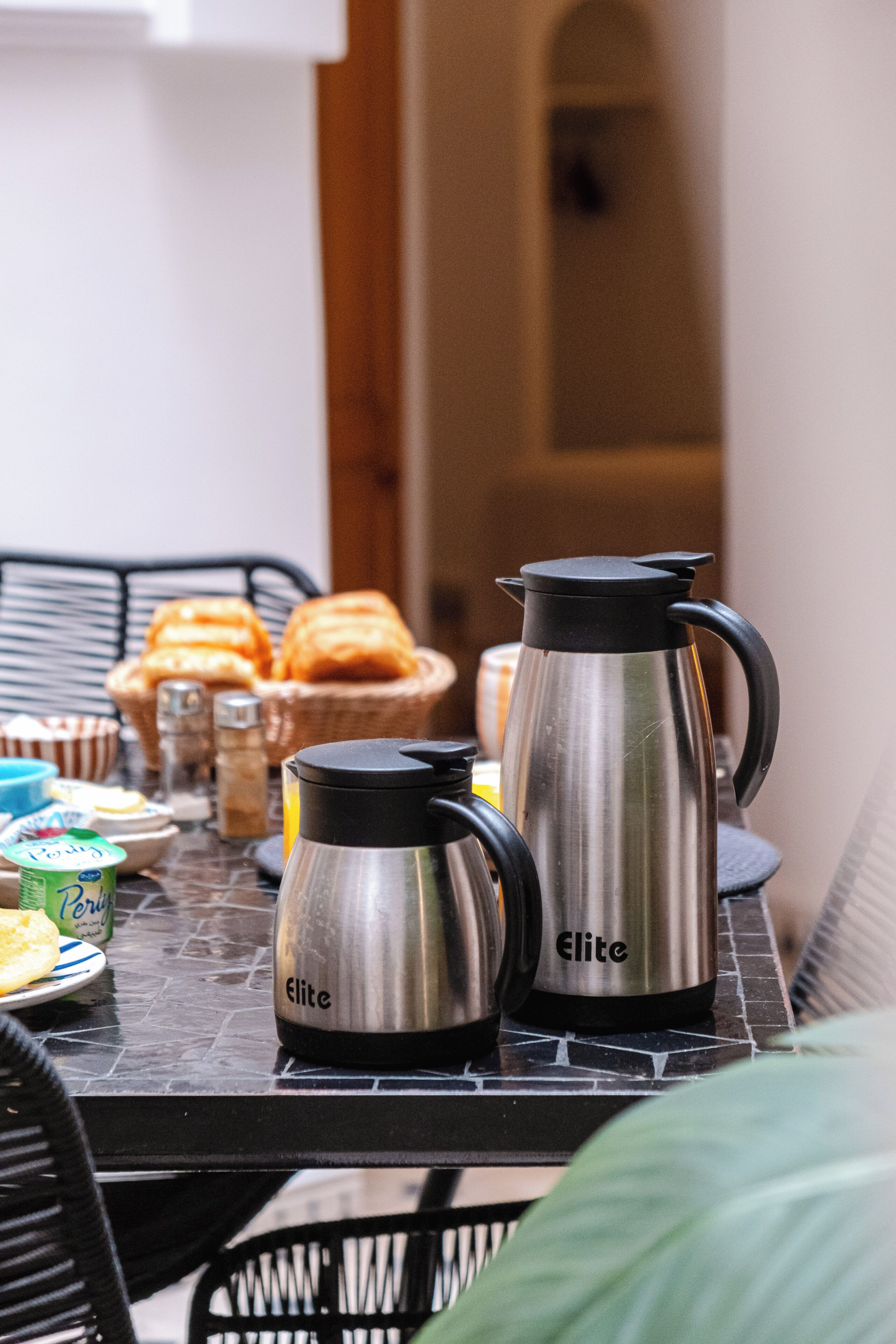 A table with two stainless steel coffee makers, a basket of bread, and various breakfast items.