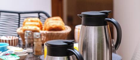 A table with two stainless steel coffee makers, a basket of bread, and various breakfast items.
