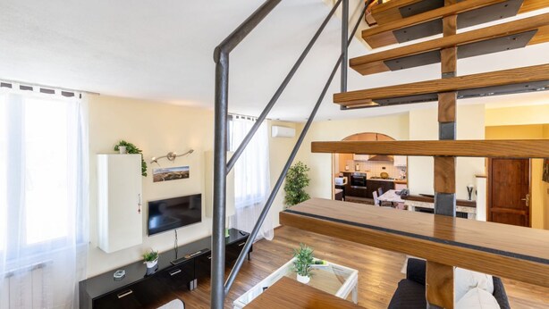 A modern living room with a wooden staircase, a flat-screen TV mounted on the wall, a dining area in the background, and a kitchen visible through an open door. - This two-level apartment located in a semi-detached house in the center of Arbia, just a 5-minute drive from Siena, is the ideal starting point for visiting the Crete Senesi, Chianti, and Val d'Orcia. Featuring a parking space, a shared garden with a pat (Arbia)