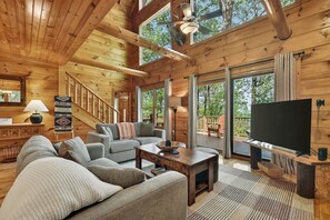 Ein Holzhäuschen im Inneren mit einer Couch, einem Couchtisch, einem Fernseher und einer Treppe, die zu einer Terrasse führt.
