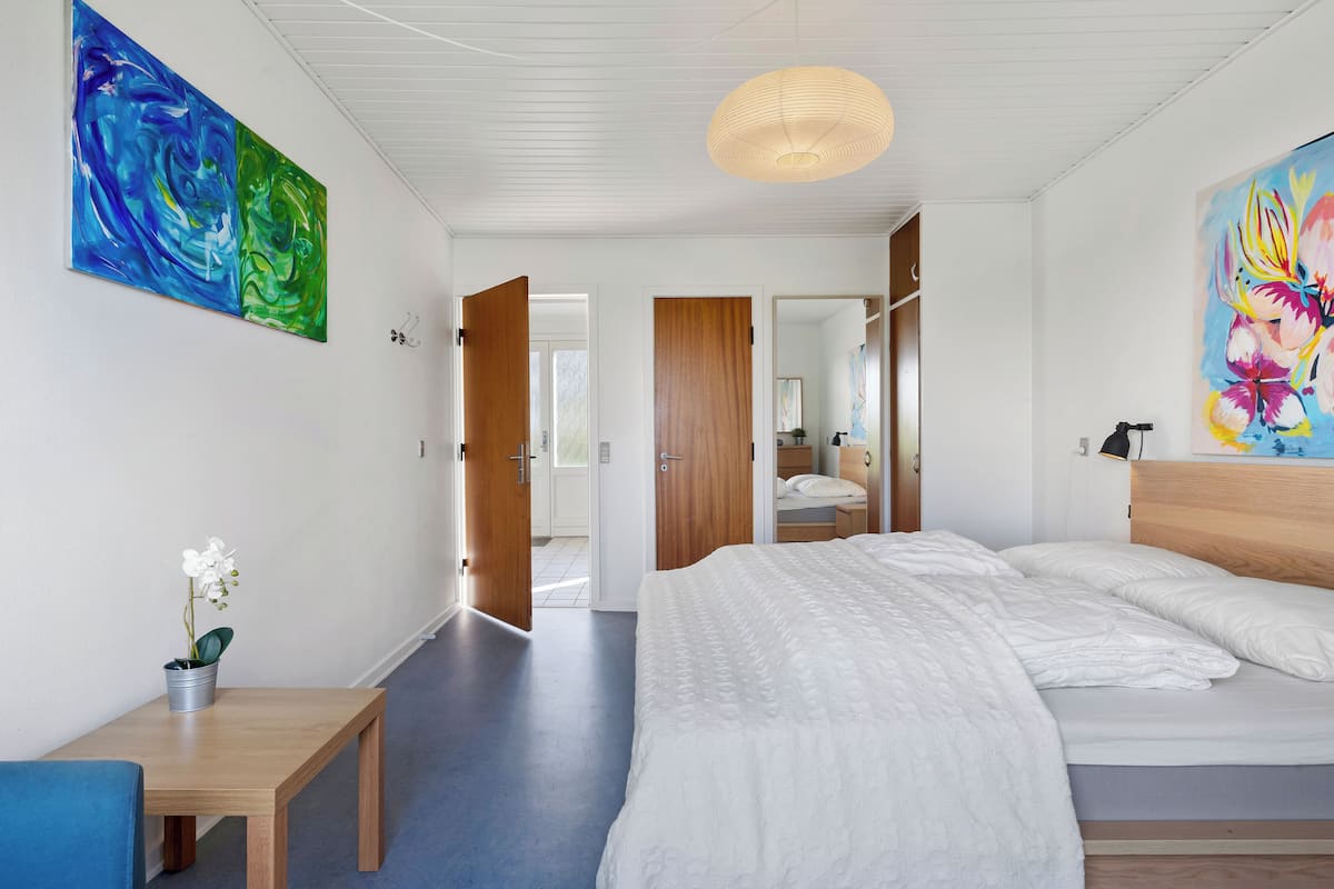 A modern bedroom with a large bed, a wooden bench, a potted orchid, and abstract paintings on the walls.