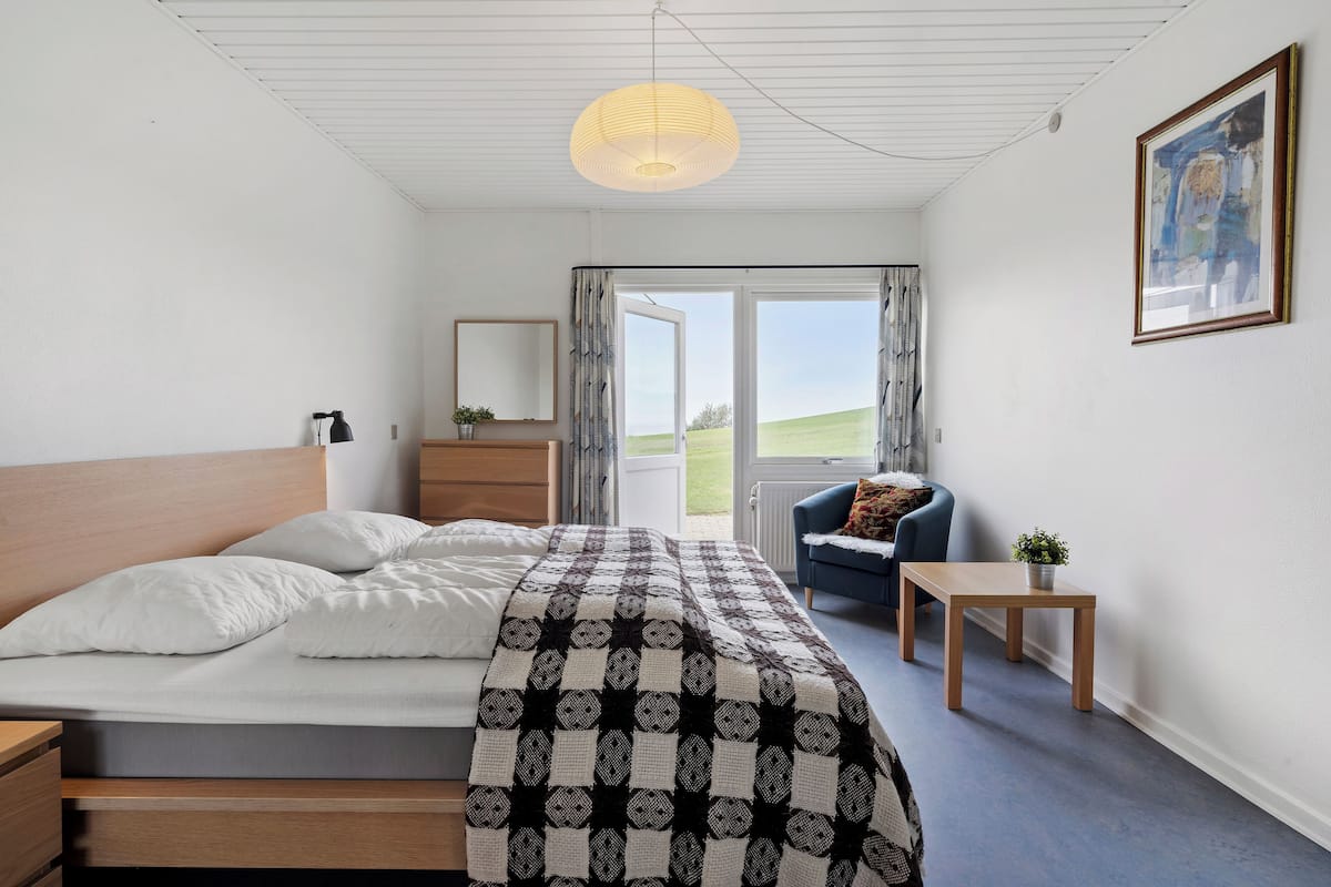 A bedroom with a bed, bedside table, a small table with a plant, a chair, and a view of the outdoors.