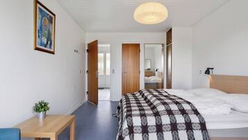 A bedroom with a bed, a bedside table with a plant, a hanging light fixture, and a door leading to another room.