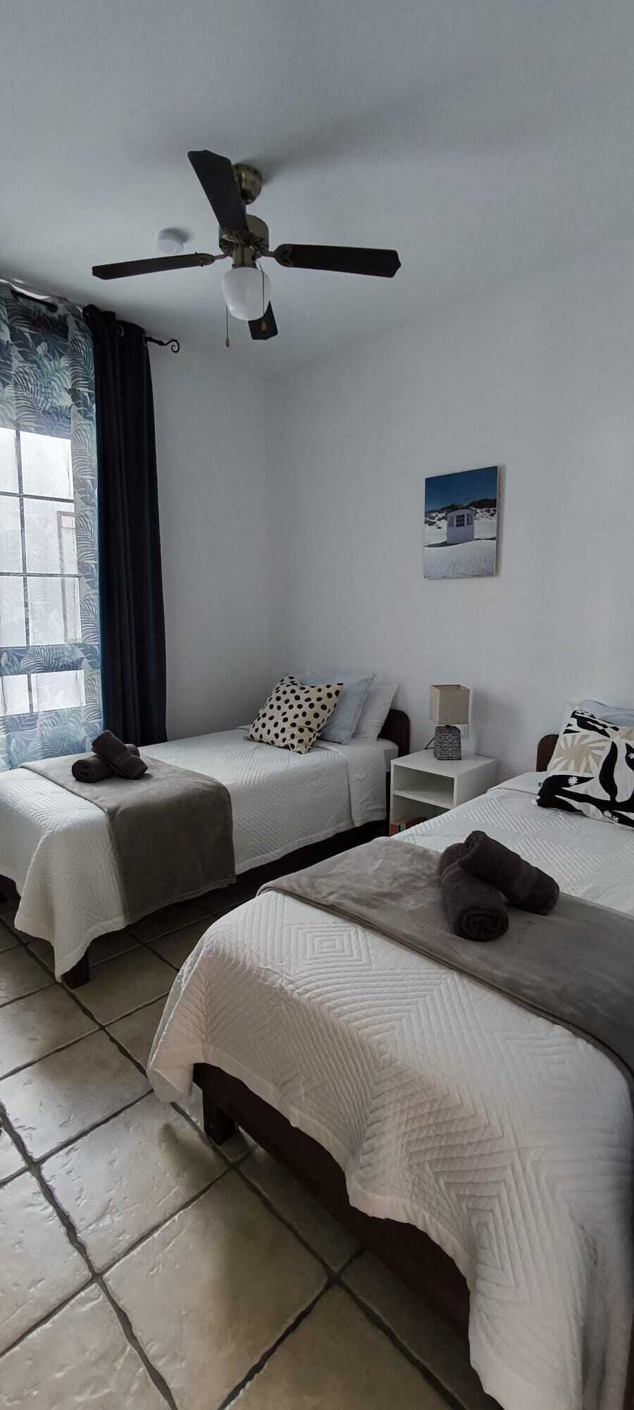 A hotel room with two beds, a ceiling fan, a window with curtains, a wall-mounted picture, and a bedside table with a lamp.