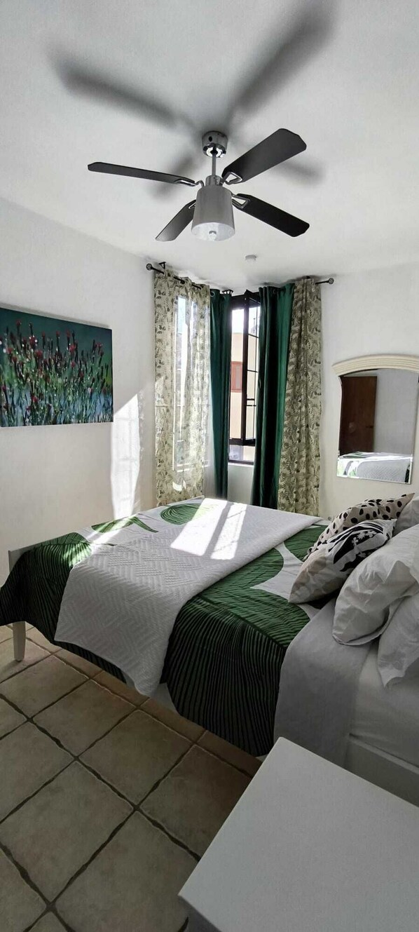 Une chambre à coucher avec un lit, un ventilateur de plafond, une fenêtre avec des rideaux et un tableau au mur.